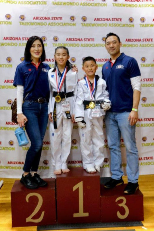 Family-of-four-standing-on-podium-at-Arizona-State-Taekwondo-Championship-two-children-in-taekwondo-uniforms-wearing-medals-parents-wearing-blue-shirts