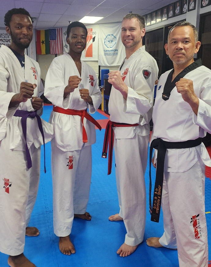 Four taekwondo students, two with black belts, one with a red belt, and one with a purple belt, posing in their uniforms inside a dojo with flags and martial arts posters on the walls.