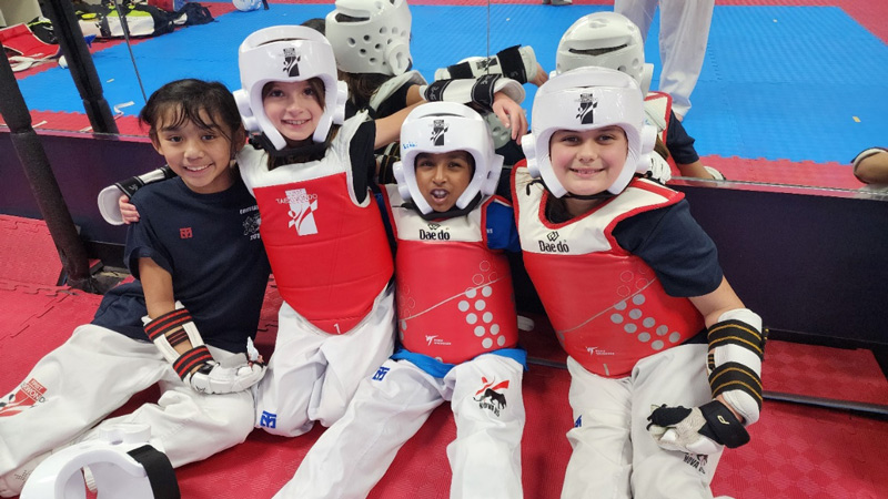 Four young girls in taekwondo sparring gear, including helmets, chest protectors, and gloves, sitting together and smiling on a red mat at the gym with a blue mat and equipment in the background.