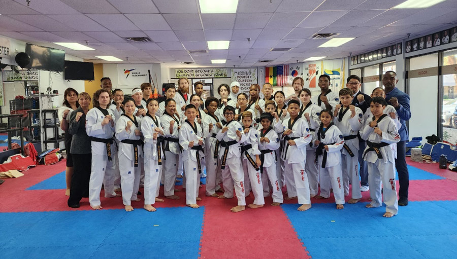 Large group of taekwondo practitioners, including both children and adults, posing in their uniforms with black belts in a dojang. The setting includes mats, banners, and training equipment in the background.
