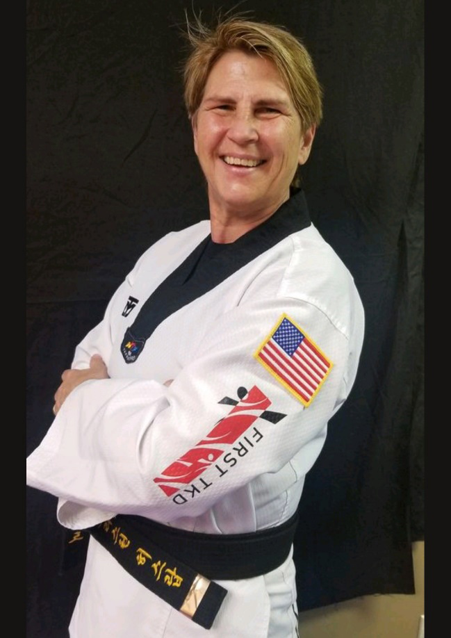Taekwondo master with a black belt and white uniform, featuring an American flag patch on the sleeve, smiling and standing against a black background.
