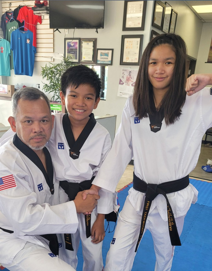 A taekwondo master kneeling beside two students, a boy and a girl, all wearing black belts and white uniforms, posing inside a dojo with framed certificates and martial arts gear displayed on the walls.
