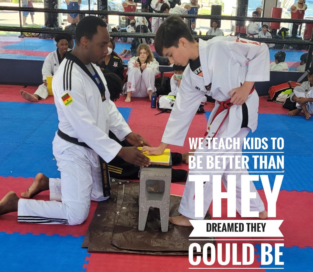 Taekwondo instructor kneeling and holding a yellow board for a young student practicing a board break in a gym. Other students in taekwondo uniforms watch in the background, with the text "We teach kids to be better than they dreamed they could be" overlaid on the image.