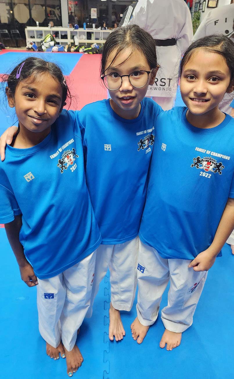 Three young girls in blue taekwondo uniforms smiling and posing together at a gym, with a colorful mat and training equipment in the background.