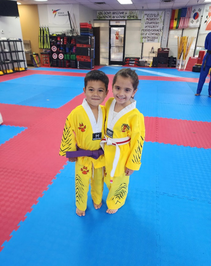 Two young taekwondo students, a boy and a girl, wearing yellow uniforms and smiling while standing on a red and blue mat inside a dojo with martial arts equipment and motivational signs in the background.